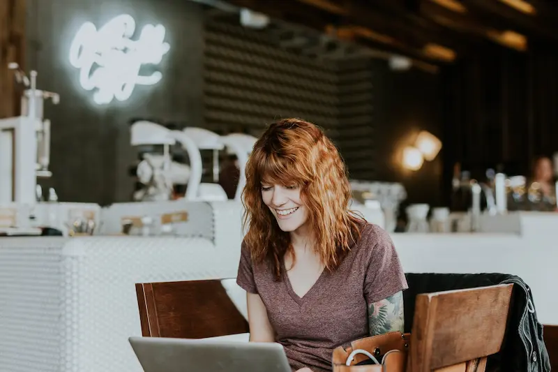 A woman with shoulder-length curly hair smiles while working on a laptop at a café table. The atmosphere appears cozy and casual, with soft lighting and blurred background elements suggesting a comfortable, inviting space. The woman's engagement with her laptop, combined with the café setting, evokes a sense of relaxed productivity.