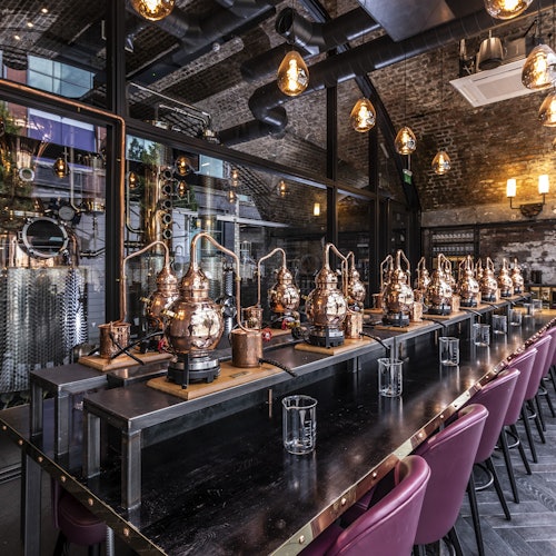 A rustic distillery with several copper stills lined up on a long table, surrounded by red barstools and industrial decor.