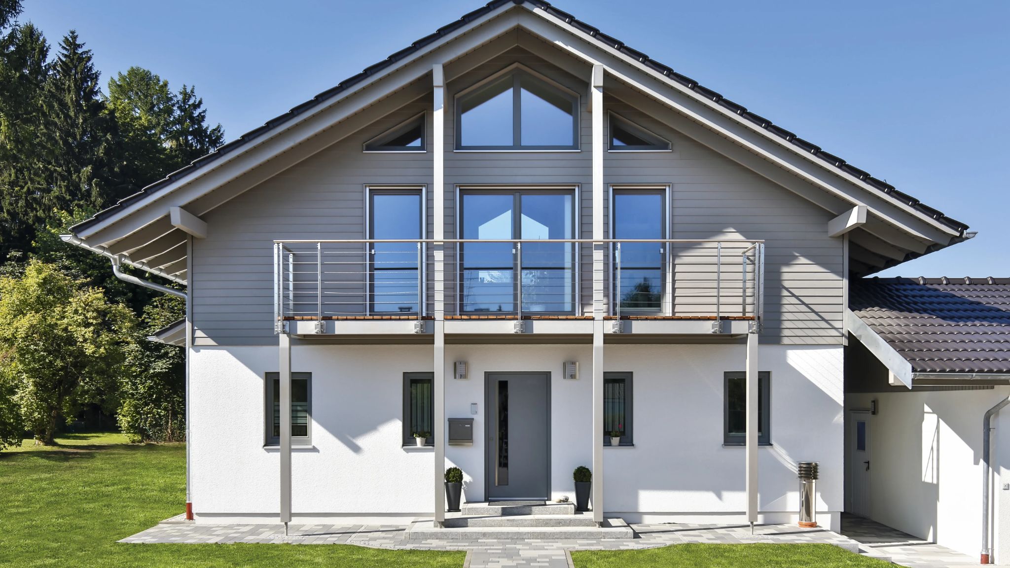 Moderne weiße Villa mit Balkon, großen Fenstern, einem schiefergrauen Satteldach und einem angelegten Garten.