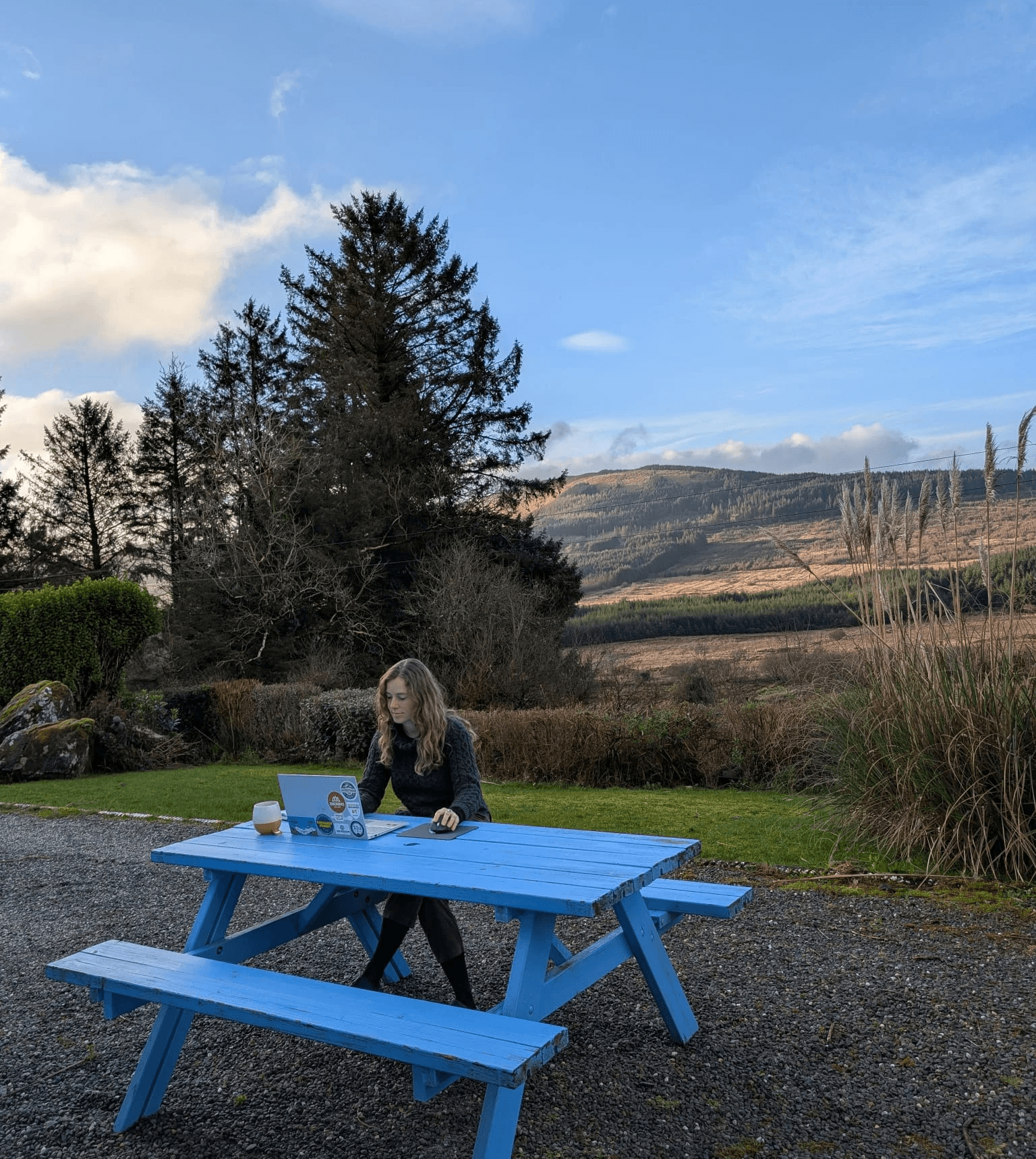 Peak Pixel Design founder Yosta working on a laptop at a blue picnic table in the Kenmare countryside, illustrating The Digital Foyer landing page service.