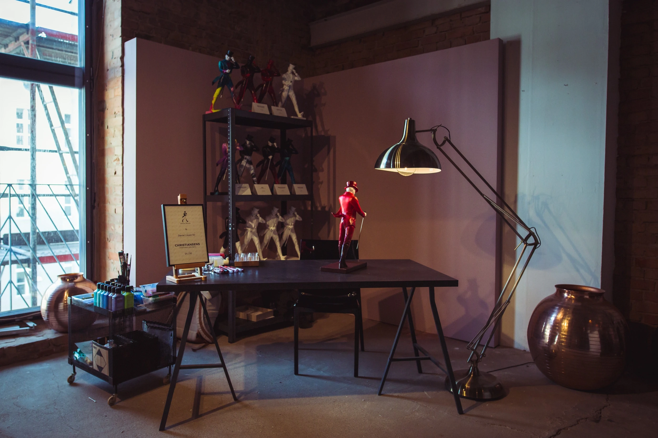 Atelier-style corner at DIAGEO BCB18 with a black work table, oversized brass architect lamp, shelf with figurines and a red sculpture against a mauve wall.