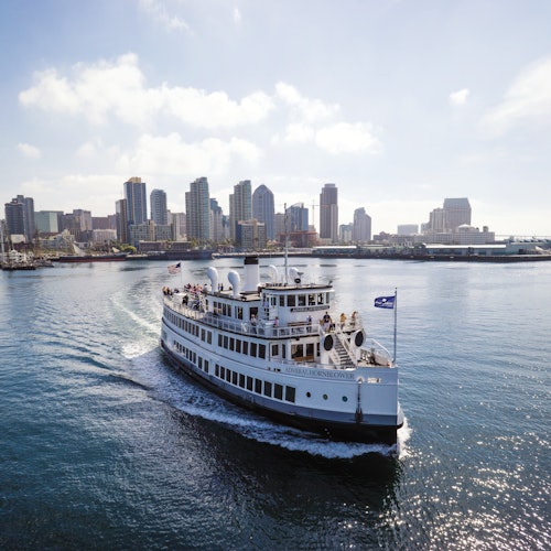 Un crucero blanco navega por un cuerpo de agua tranquila con un horizonte de la ciudad lleno de edificios altos al fondo bajo un cielo parcialmente nublado.