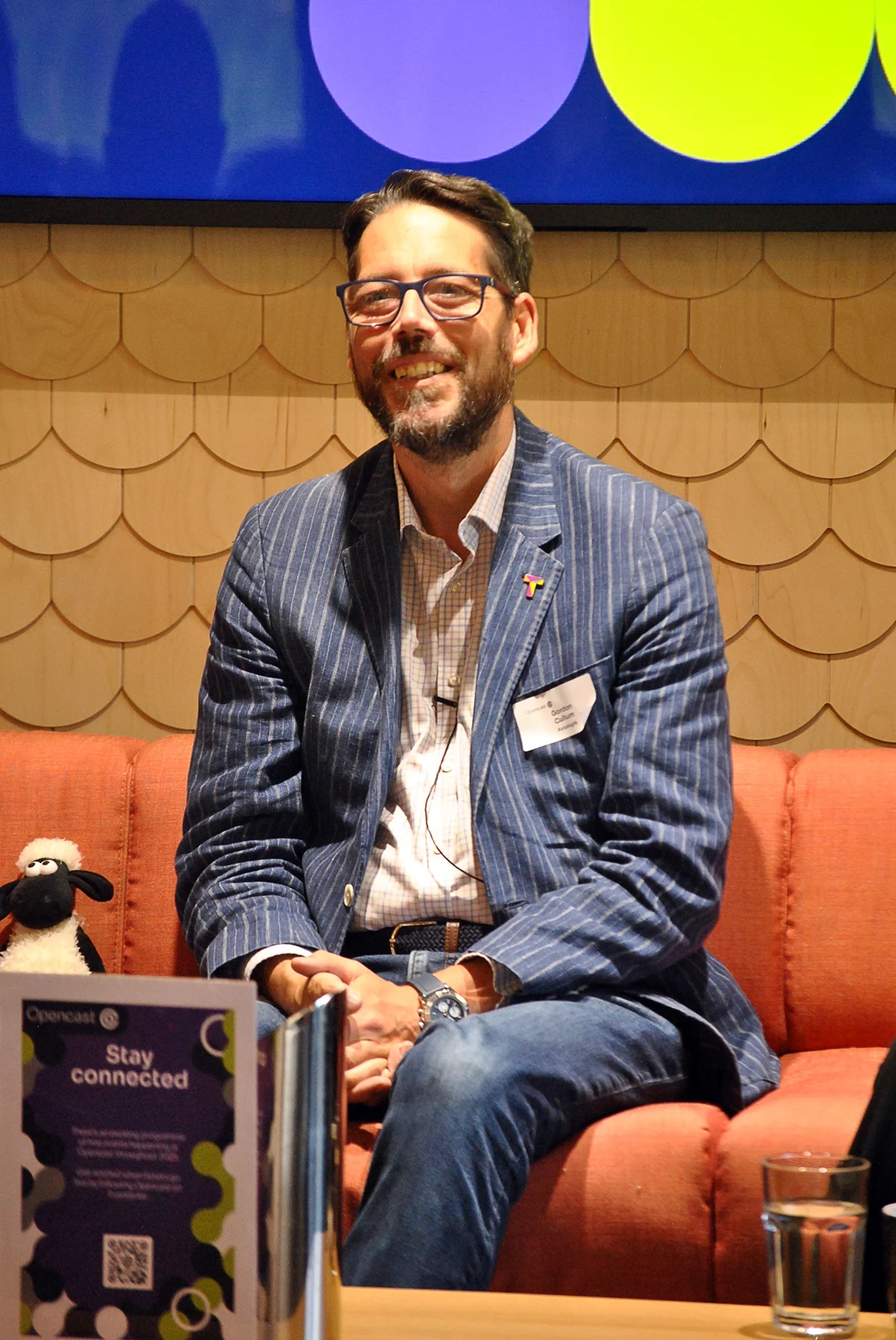 Person seated on an orange sofa wearing a blue pinstripe jacket, with a brochure and glass on the table in front.