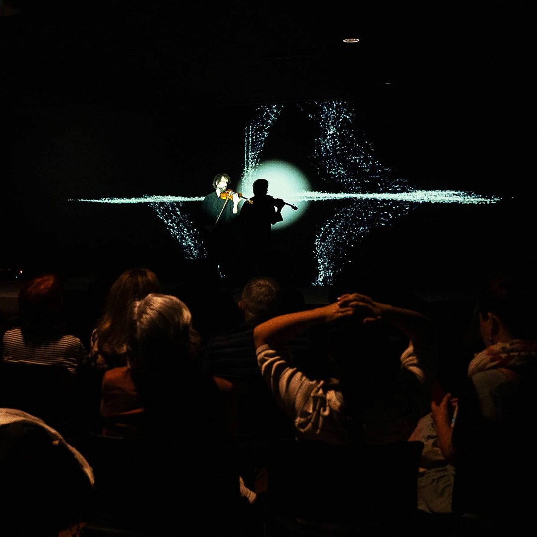 Audience in front of a man playing the violin with a projection of organic shapes and blue granular texture