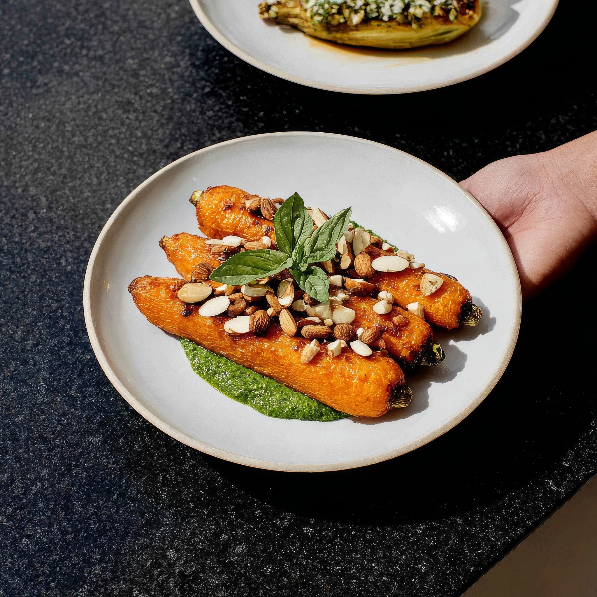 White plate with roasted carrots served on green sauce, topped with almonds and fresh herbs, held by a hand over a dark table.