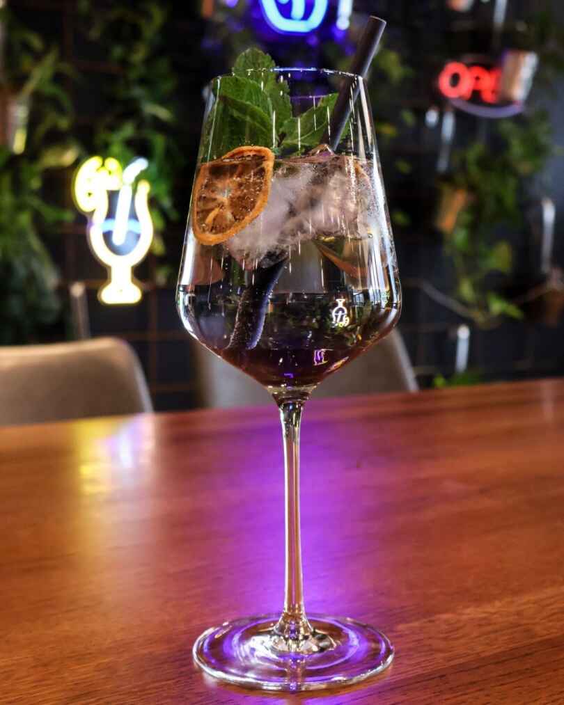 A cocktail in a wine glass garnished with dried citrus and mint, placed on a wooden table with neon lights in the background.