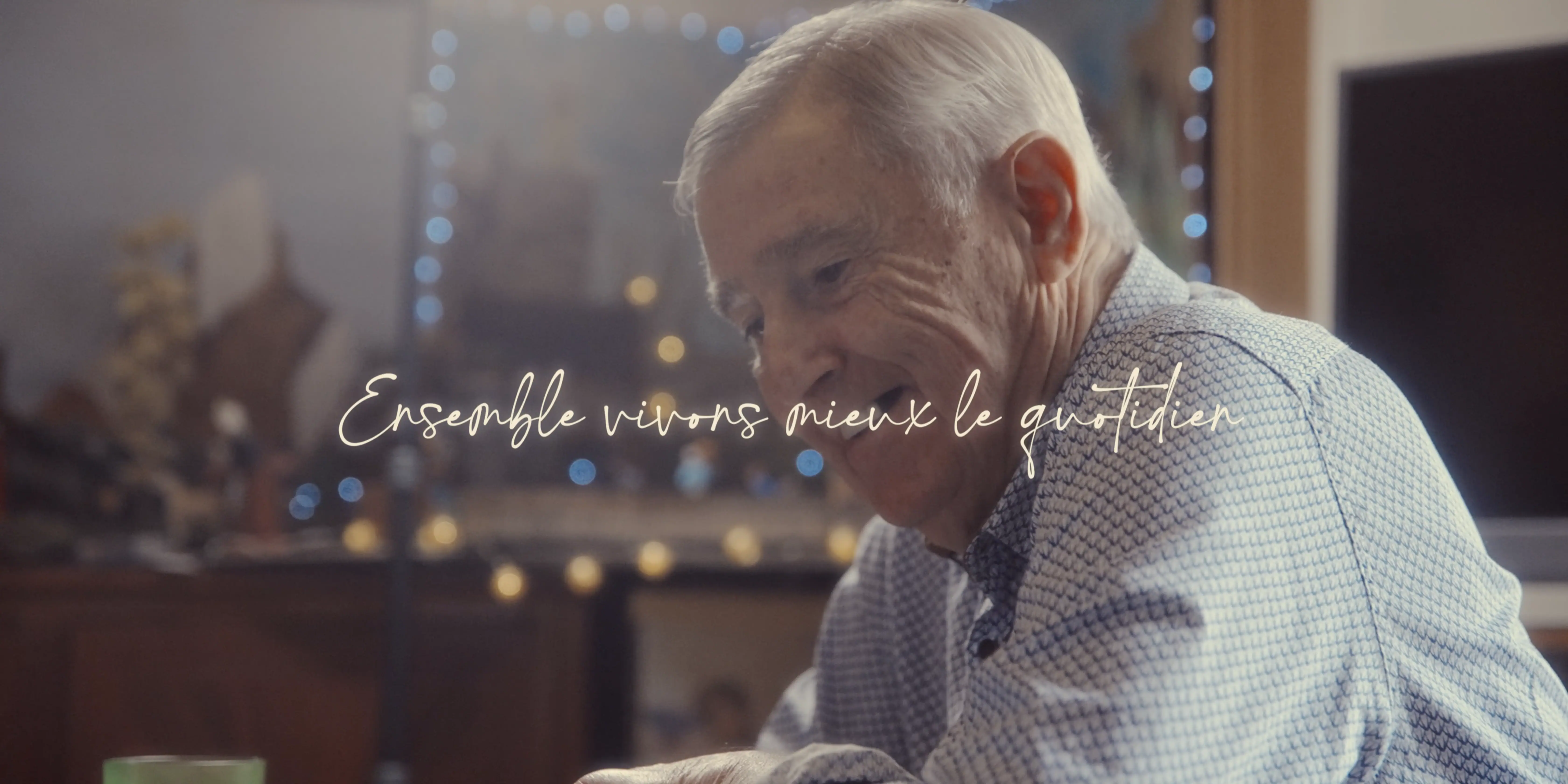 A smiling older man with gray hair is seated indoors at a table, wearing a checkered shirt, with blurred festive lights in the background and overlayed text in French.