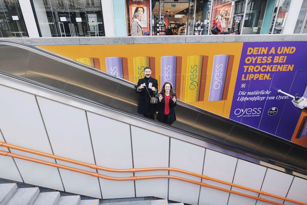 Mann und Frau auf Rolltreppe halten OYESS Produkte vor Plakat