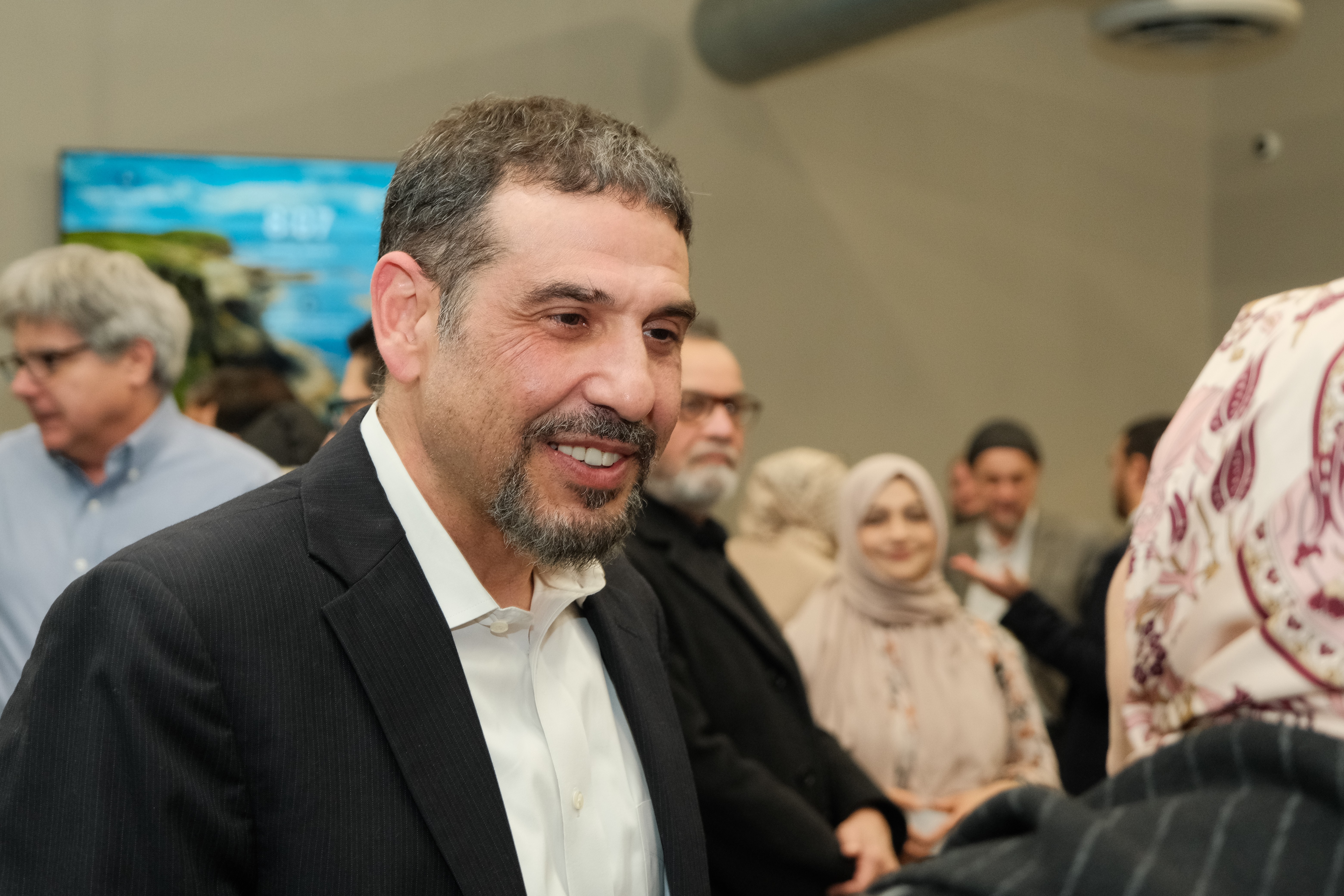 A smiling man in a suit interacts with a woman in a headscarf, amidst a diverse group of people.
