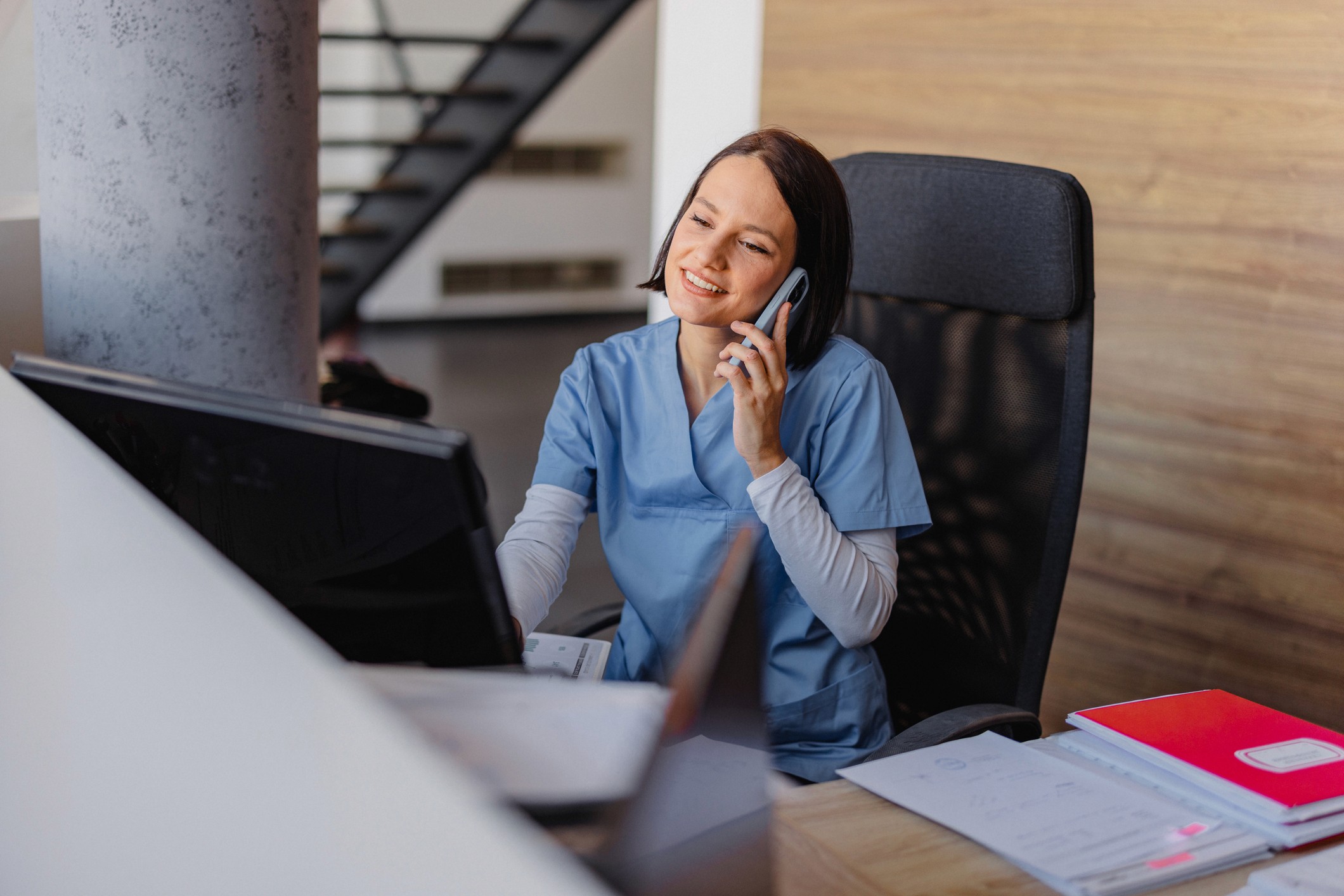 A young woman with short hair wearing medical scrubs sits in a reception area working on a computer whilst taking a phone call on her phone.