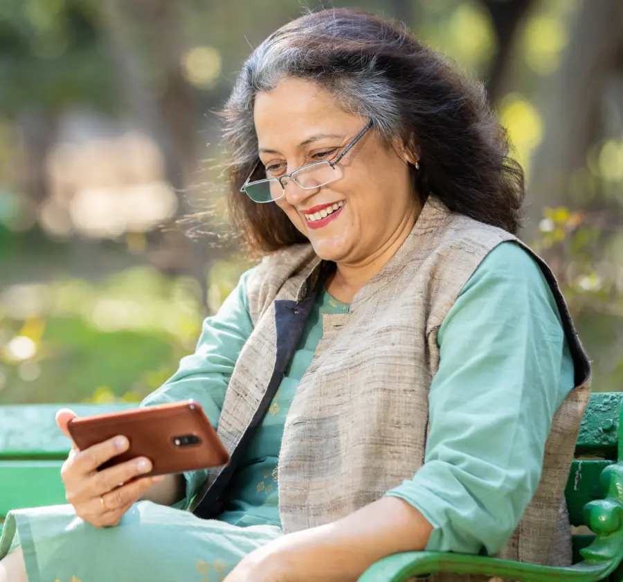 An older Indian origin woman sitting on a park bench, smiling while using her smartphone, representing the ease of access to digital health solutions for older adults managing chronic conditions like diabetes.