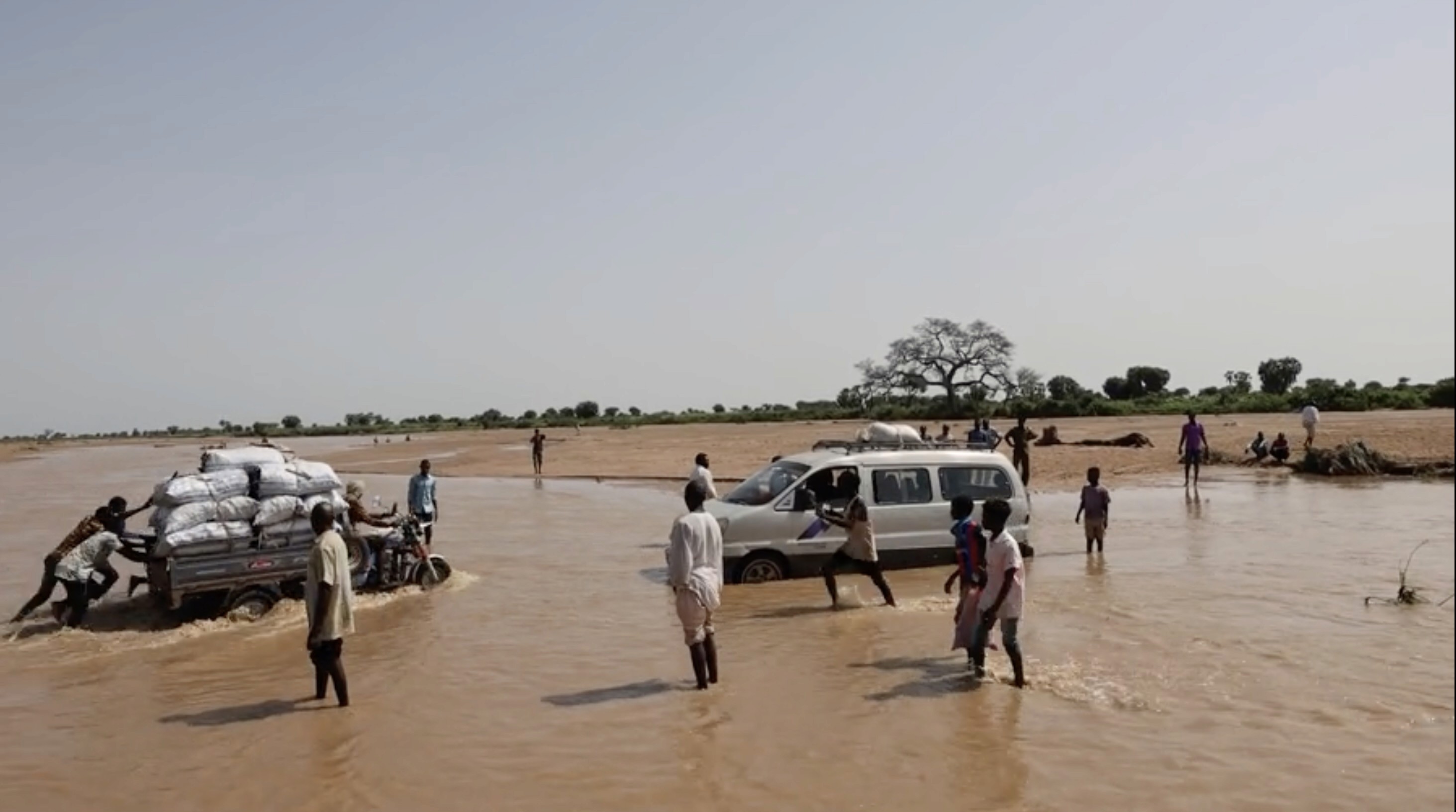 Contry side of sub-saharan Africa affected by flash floods. A goup of people is helping a moto truck to pass through a flooded area while other car is stuck in the mud.