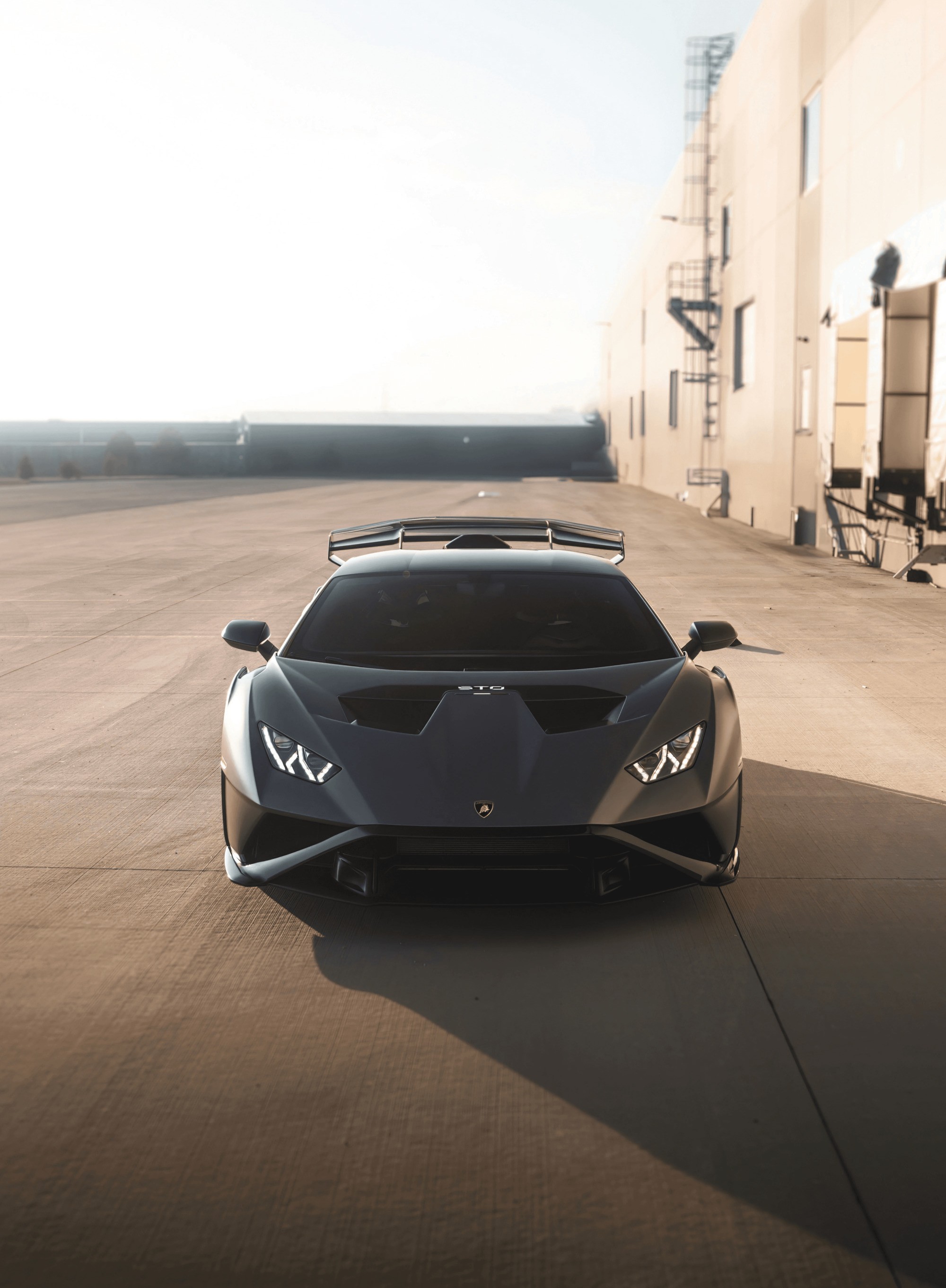 A sleek black sports car with a low profile and angular design is parked on an industrial concrete road, with a beige building and loading docks in the background under a clear sky.