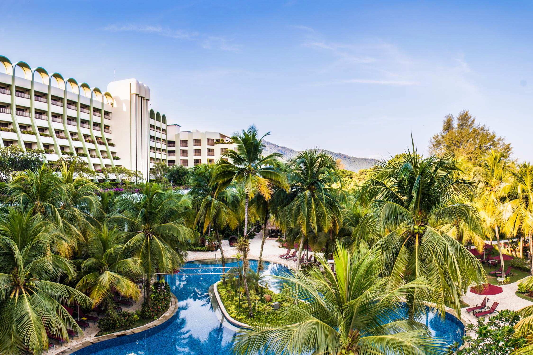 A multi-story white resort building with a curved facade overlooks a turquoise swimming pool surrounded by palm trees.