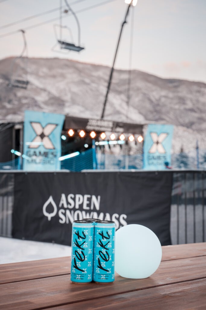 Two blue beverage cans and a glowing table light in front of X Games and Aspen Snowmass signage