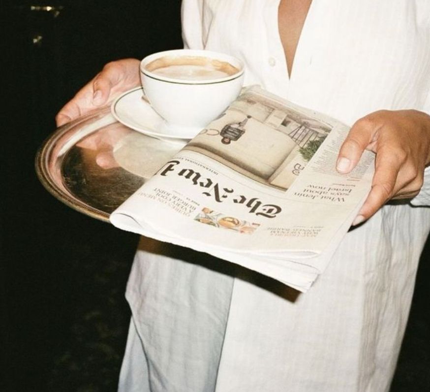 Person with a mustache wearing a black t-shirt carrying a tall stack of pizza boxes inside a dimly lit bar or restaurant.