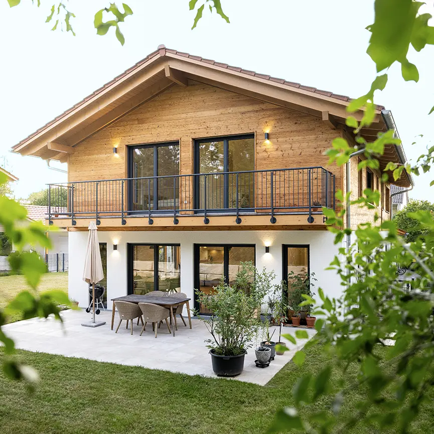 Zweistöckiges Haus mit Holzfassade im Obergeschoss, großem Balkon mit Metallgeländer und Terrasse mit Gartenmöbeln, umgeben von Grünpflanzen