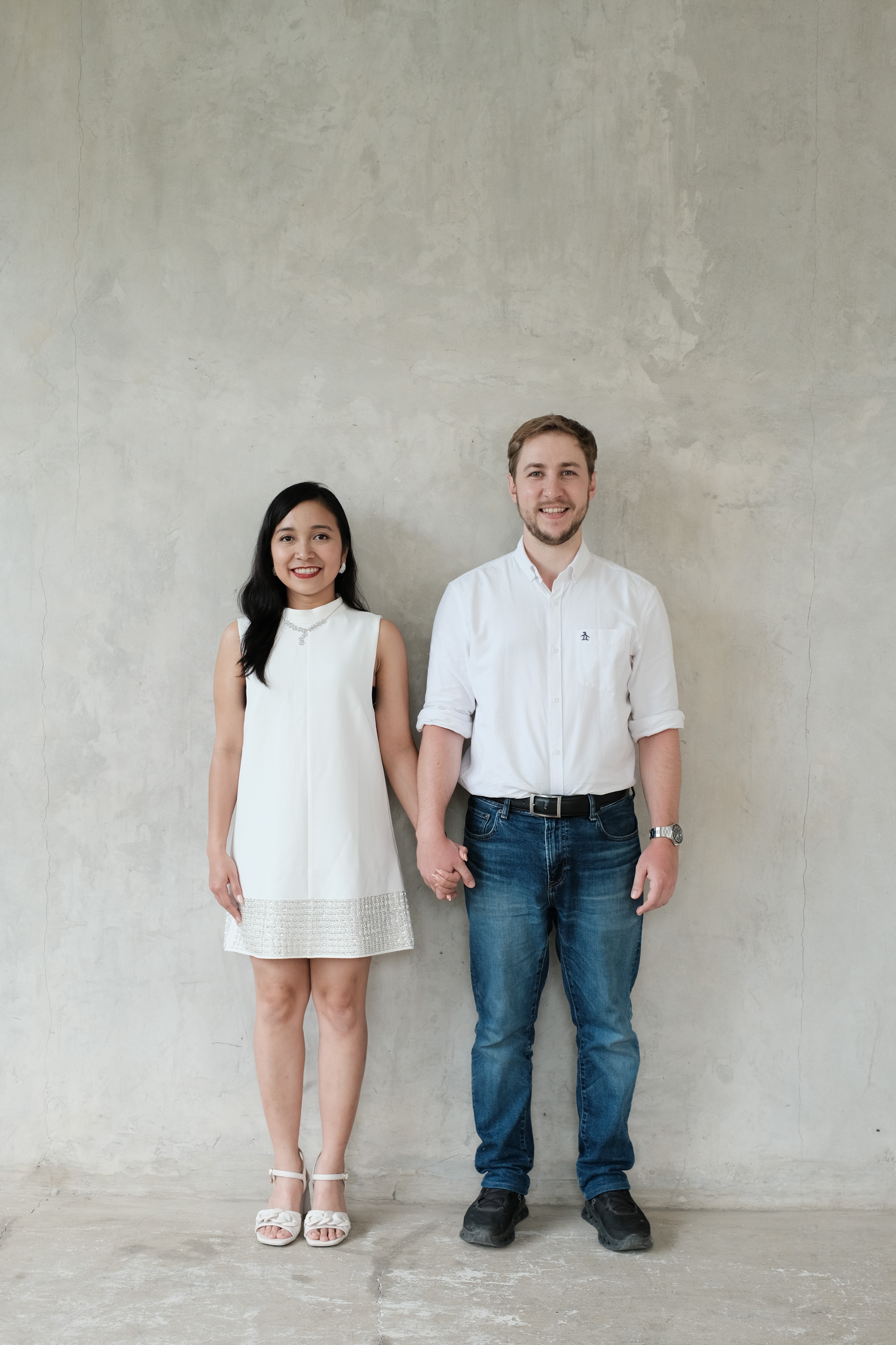 A couple stands on a sandy hill at sunset, facing away, with arms raised in a relaxed "hang loose" gesture. The scene conveys tranquility and joy.