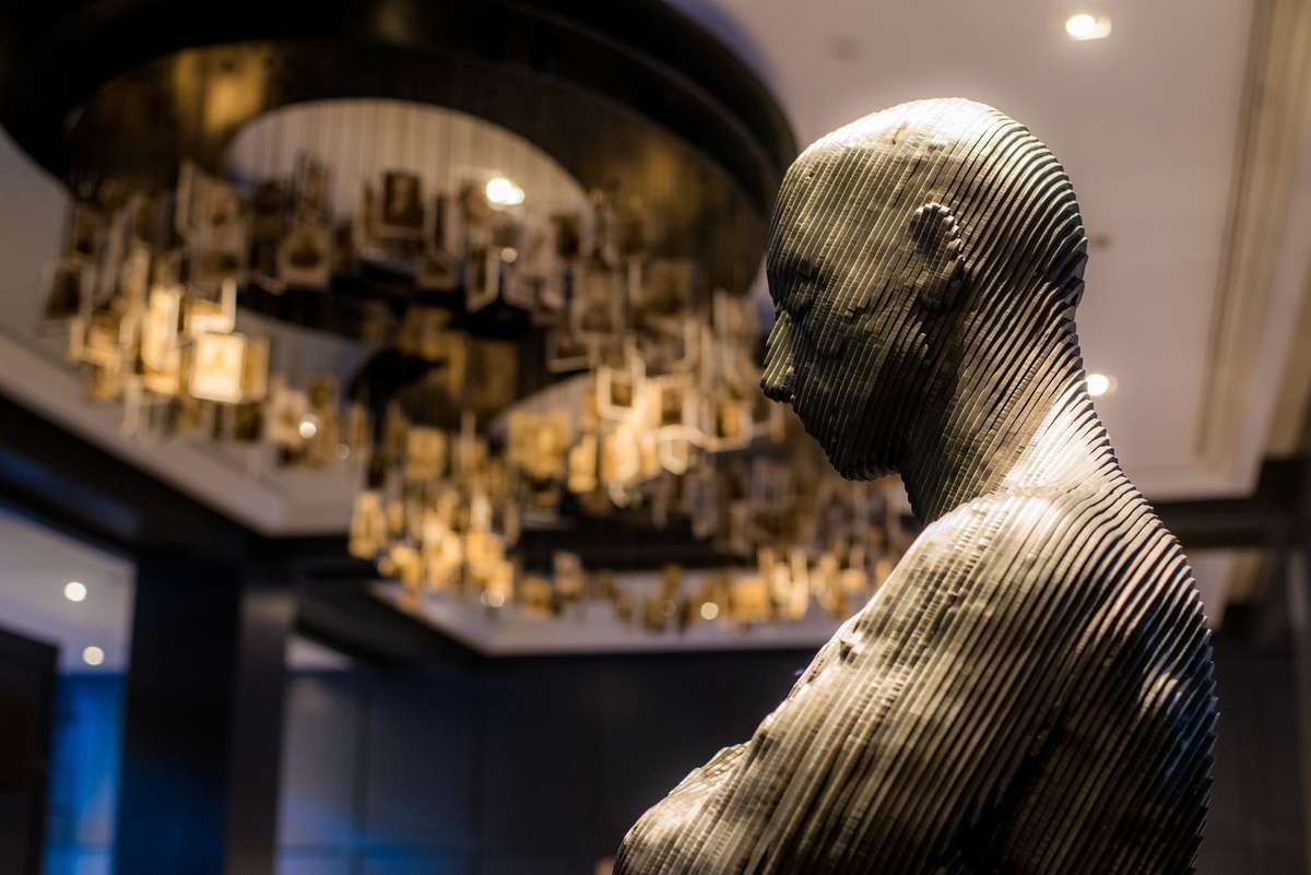 A textured bronze sculpture of a human head and torso stands in front of a large, ornate gold chandelier.