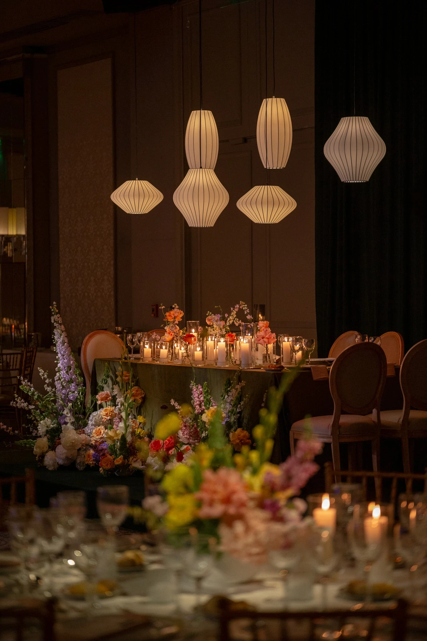 Salón de eventos con lámparas de papel plisado sobre mesas principales decoradas con flores y velas cálidas.