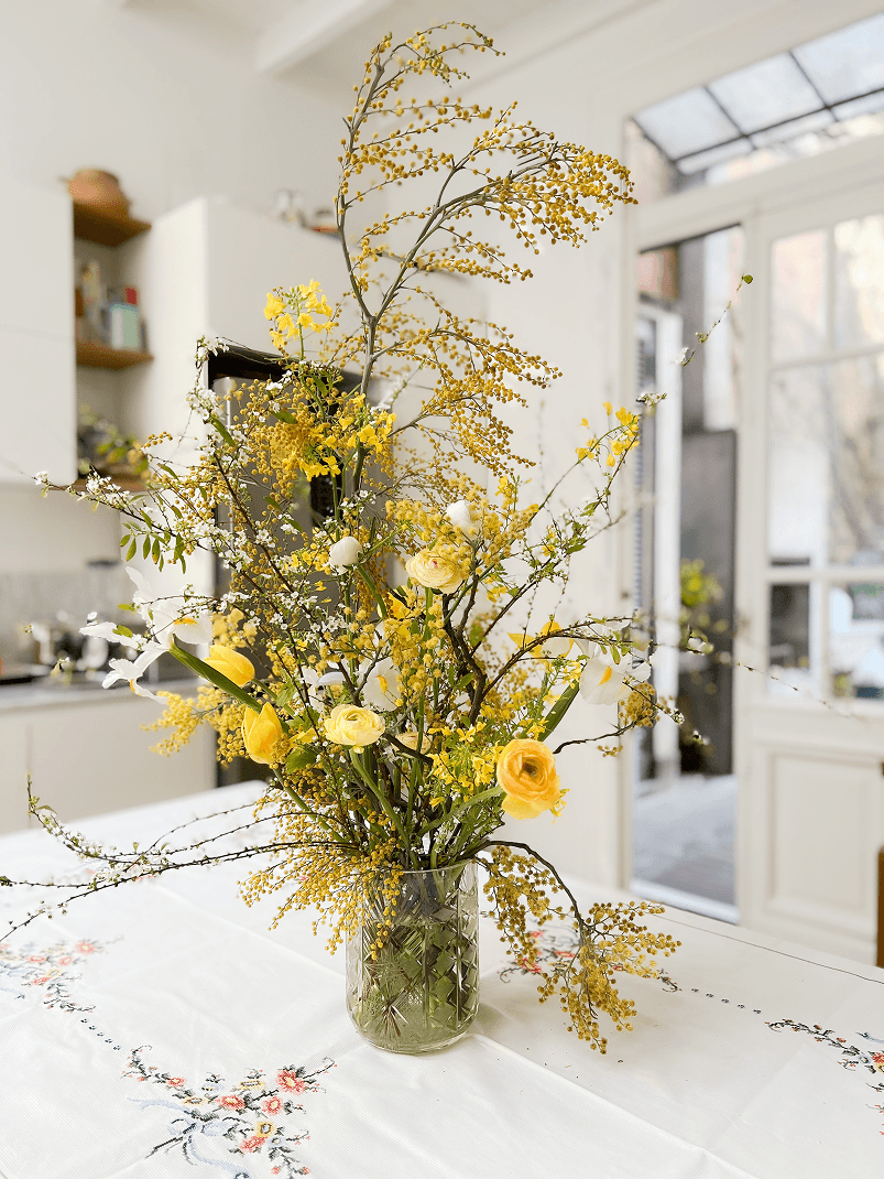 Ramo de flores amarillas y blancas con ramas verdes en un florero de vidrio, colocado sobre una mesa con mantel bordado en una cocina luminosa.