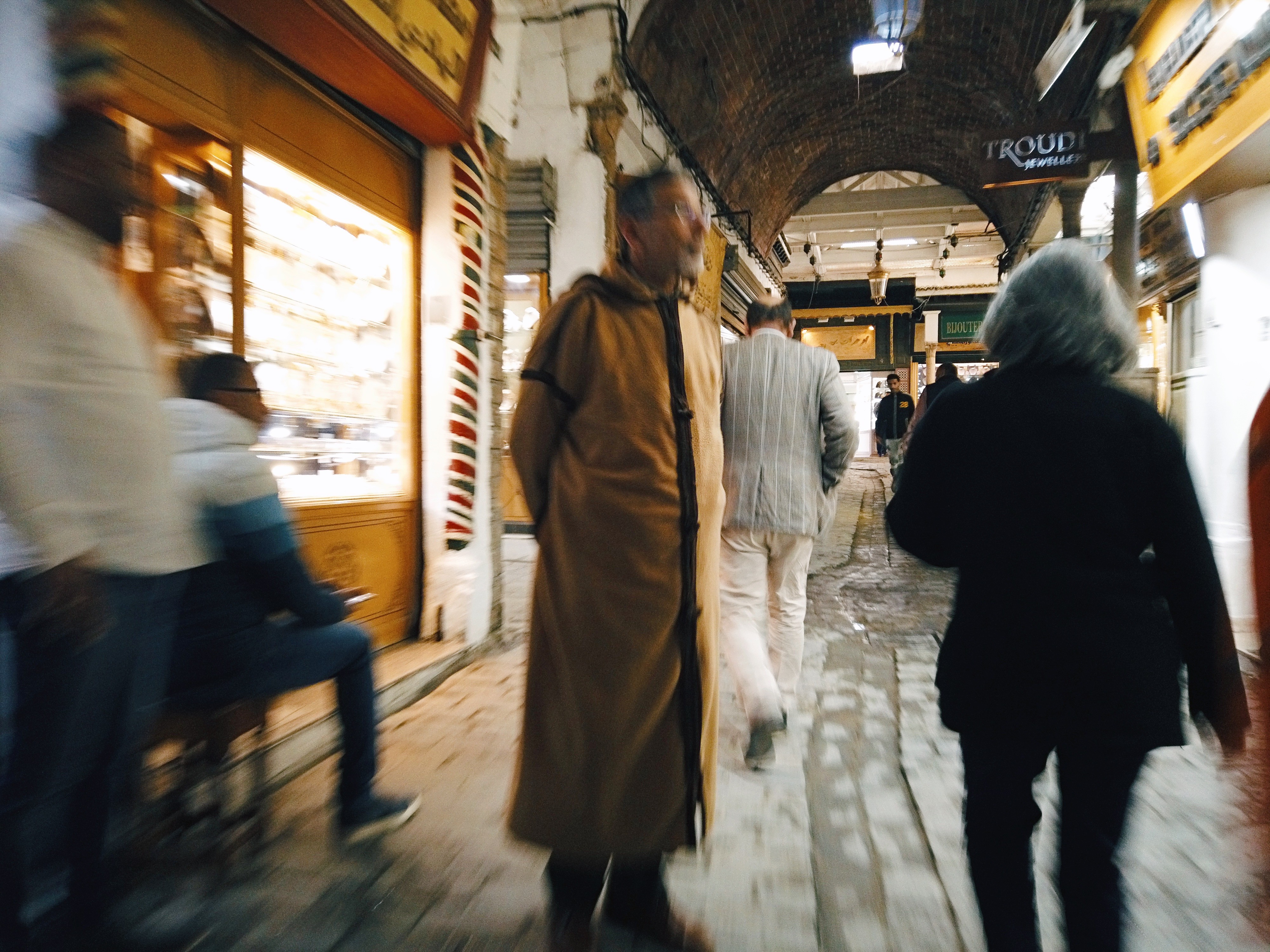 Photographie de rue d'un homme les bras dans le dos portant une robe traditionnelle se tenant au mlieu d'une artère du souk de Tunis