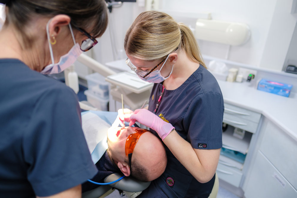 Two dental professionals in scrubs and masks perform a procedure on a patient in a modern dental office.