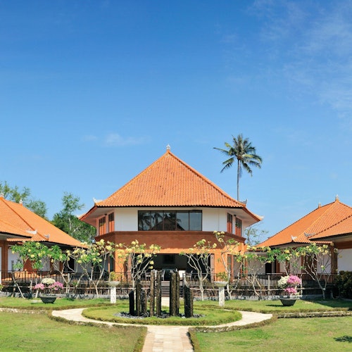 一座大型橙色屋顶的建筑,周围有较小的结构、翠绿的植被,背景是一片晴朗的蓝天。