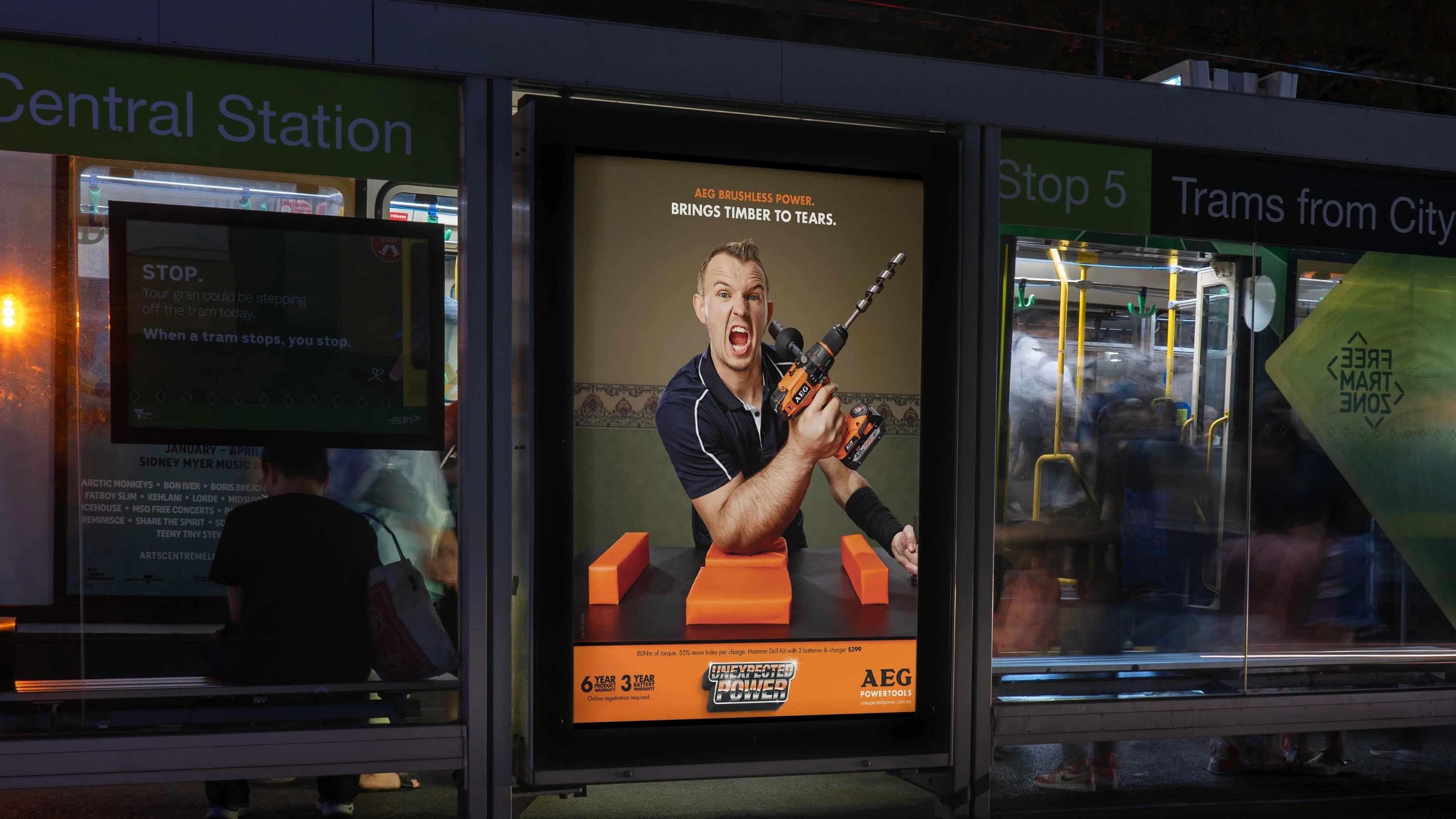 AEG bus shelter poster: 'Brings timber to tears.' with a man holding a drill on orange blocks at a tram stop