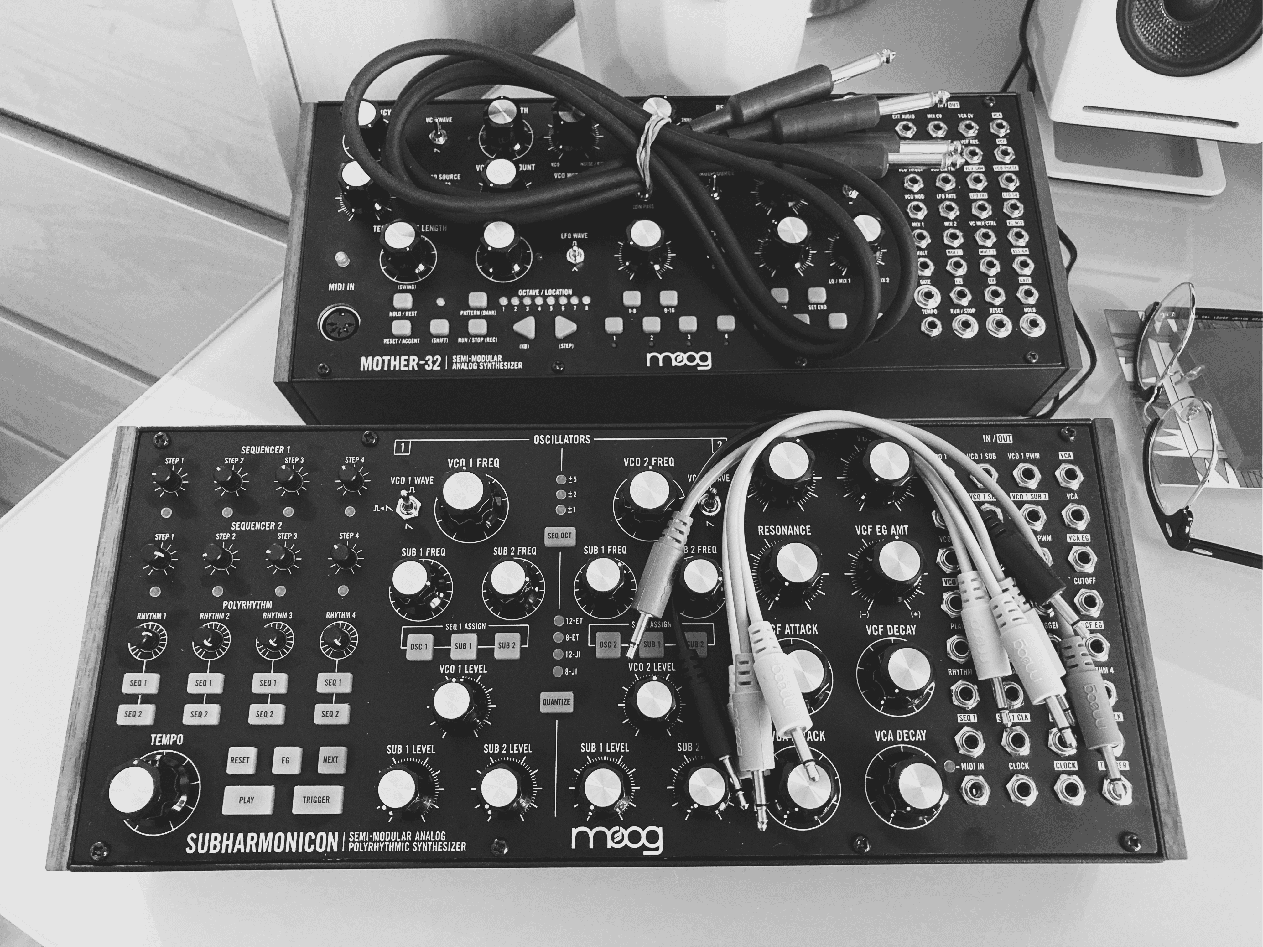 ‘Black-and-white photograph of two Moog semi-modular synthesizers on a tabletop: the top unit is a Mother-32 with coiled patch cables draped over its array of knobs and jacks, and the bottom unit is a Subharmonicon featuring sequencer knobs, oscillator and filter dials, and multiple patch cables plugged into its panels; a pair of eyeglasses and a speaker appear at the right edge.’