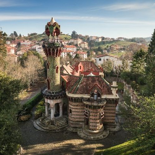 An ornate, multi-colored building with a tower, surrounded by greenery, with a town and hills in the background under a blue sky.