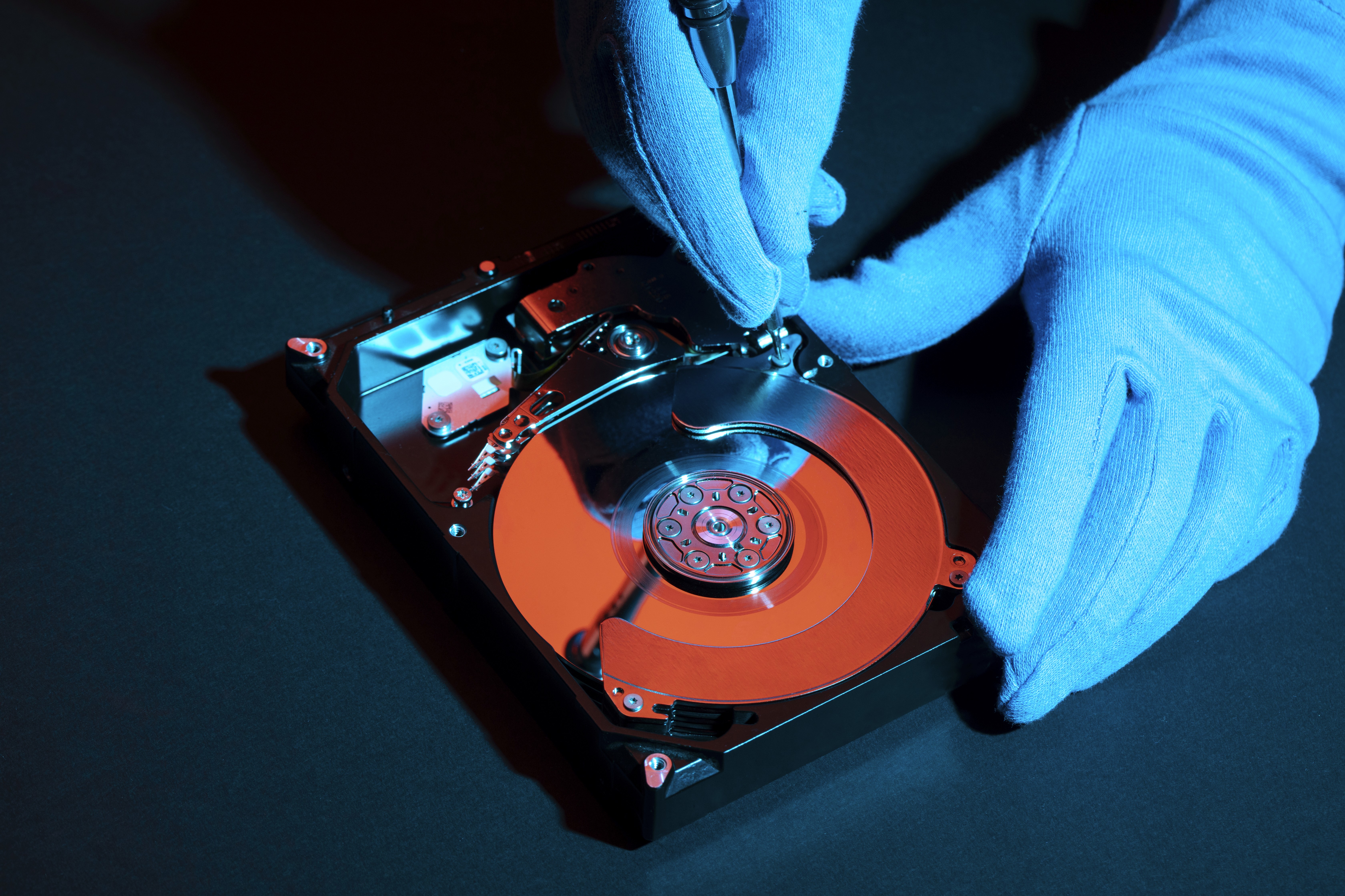 A person wearing blue anti-static gloves uses a screwdriver to repair or inspect an open hard disk drive under blue and red lighting, showing the exposed platters and actuator arm.