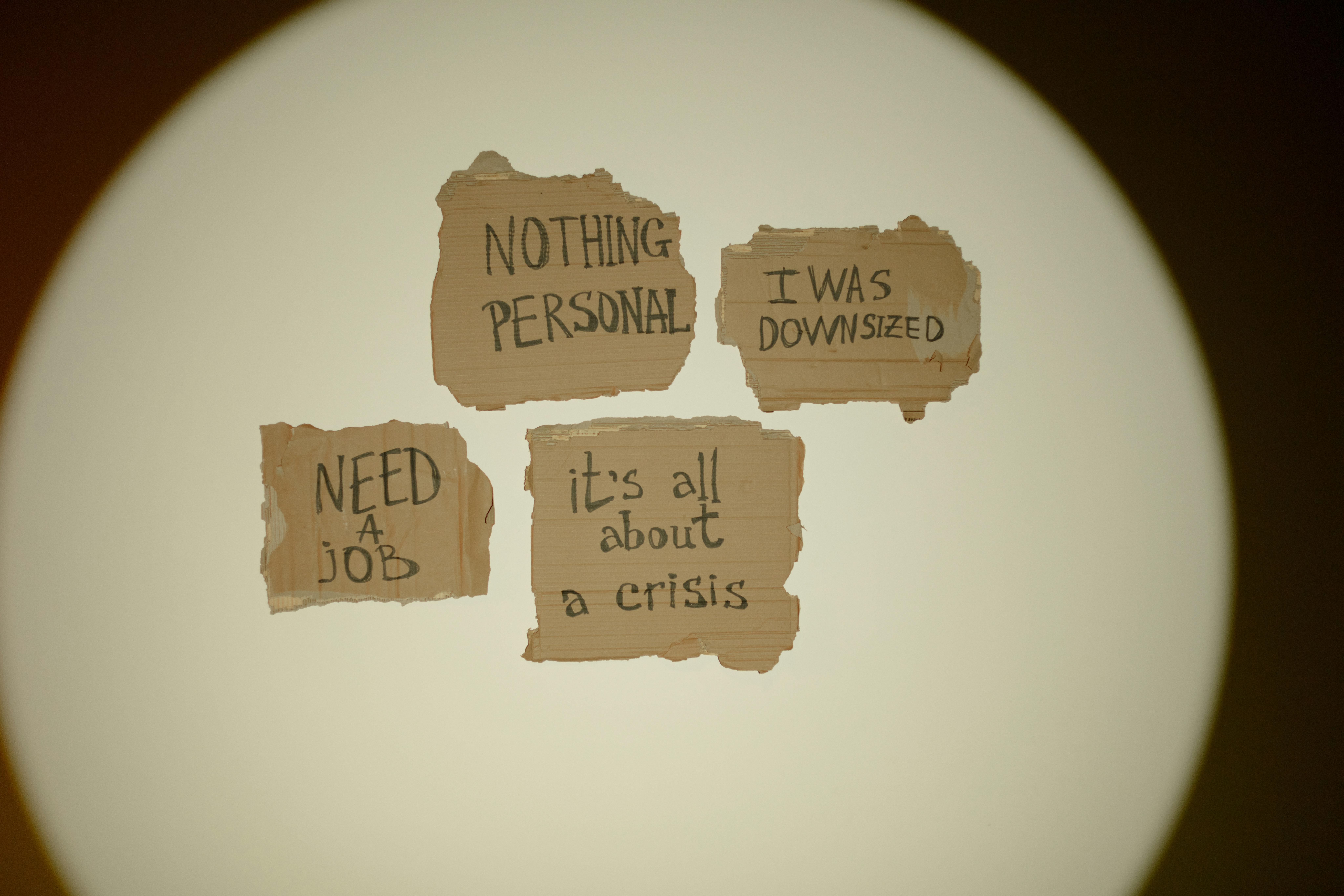 Cartoncini con frasi scritte a mano come ‘I was downsized’ e ‘Need a job’, rappresentazione simbolica di crisi occupazionale e perdita del lavoro.
