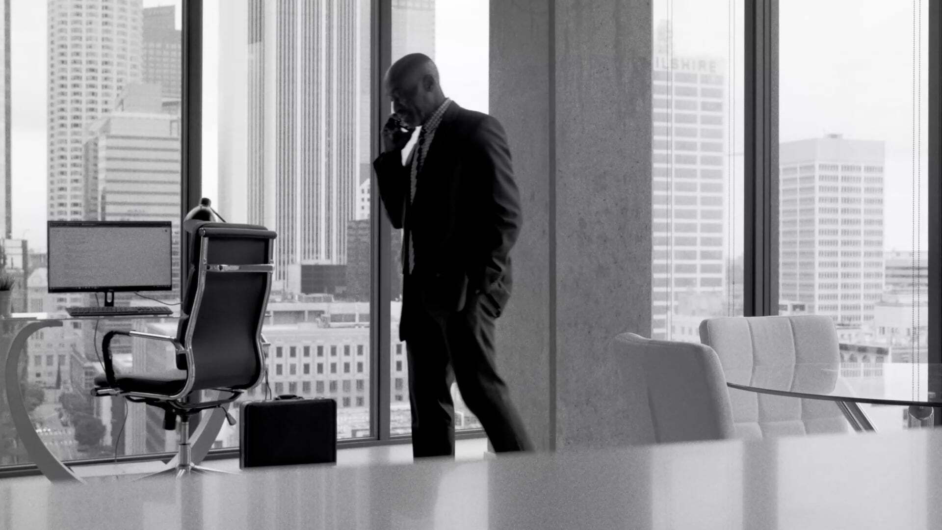 A black and white image of a man in a suit speaking on the phone in a modern office setting