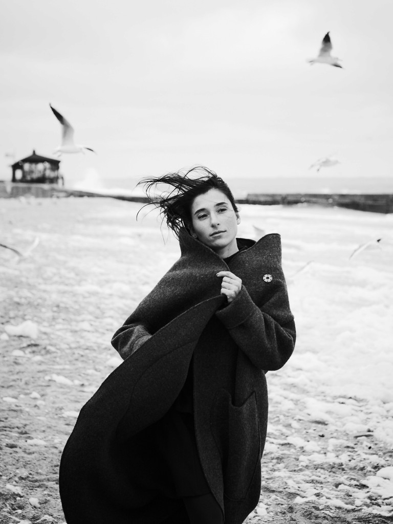 black white image showing a woman on a beach with seagulls flying behind her.