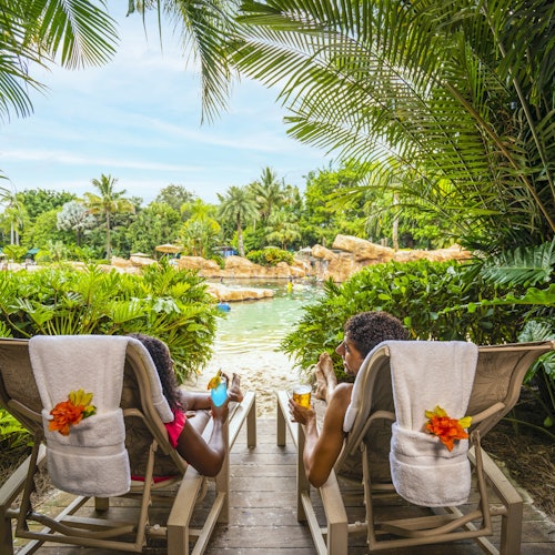 Двама души се отпускат на столове с напитки, заобиколени от тропически растения, гледайки към богата зелена гледка с камъни и вода.