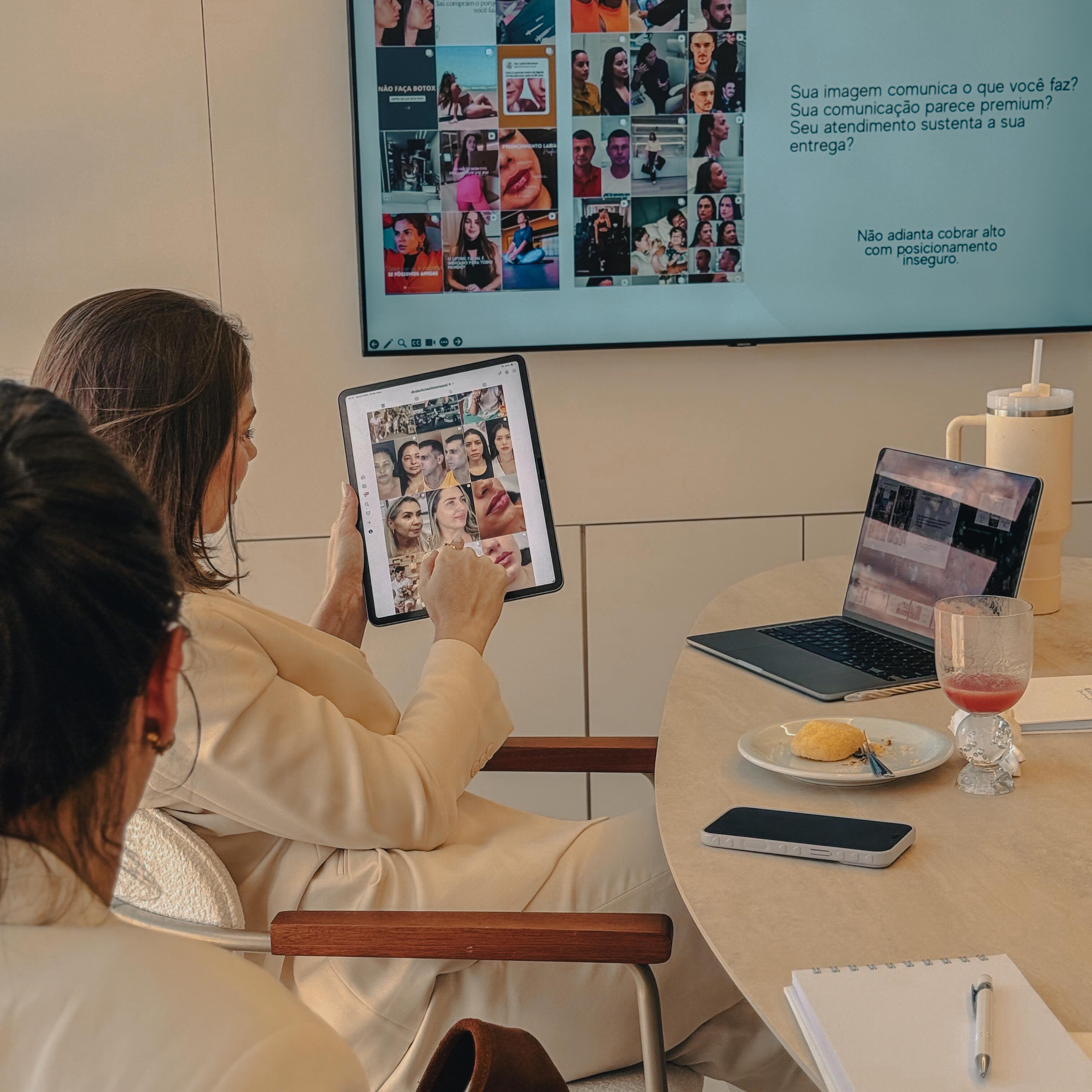 Consultora segurando tablet com grade de imagens de redes sociais, com o mesmo conteúdo projetado na TV da sala