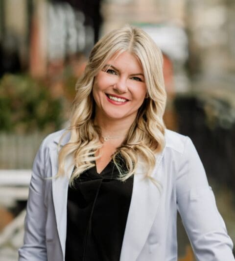 A woman with long, wavy blonde hair smiles confidently while wearing a black top and light blazer outdoors.