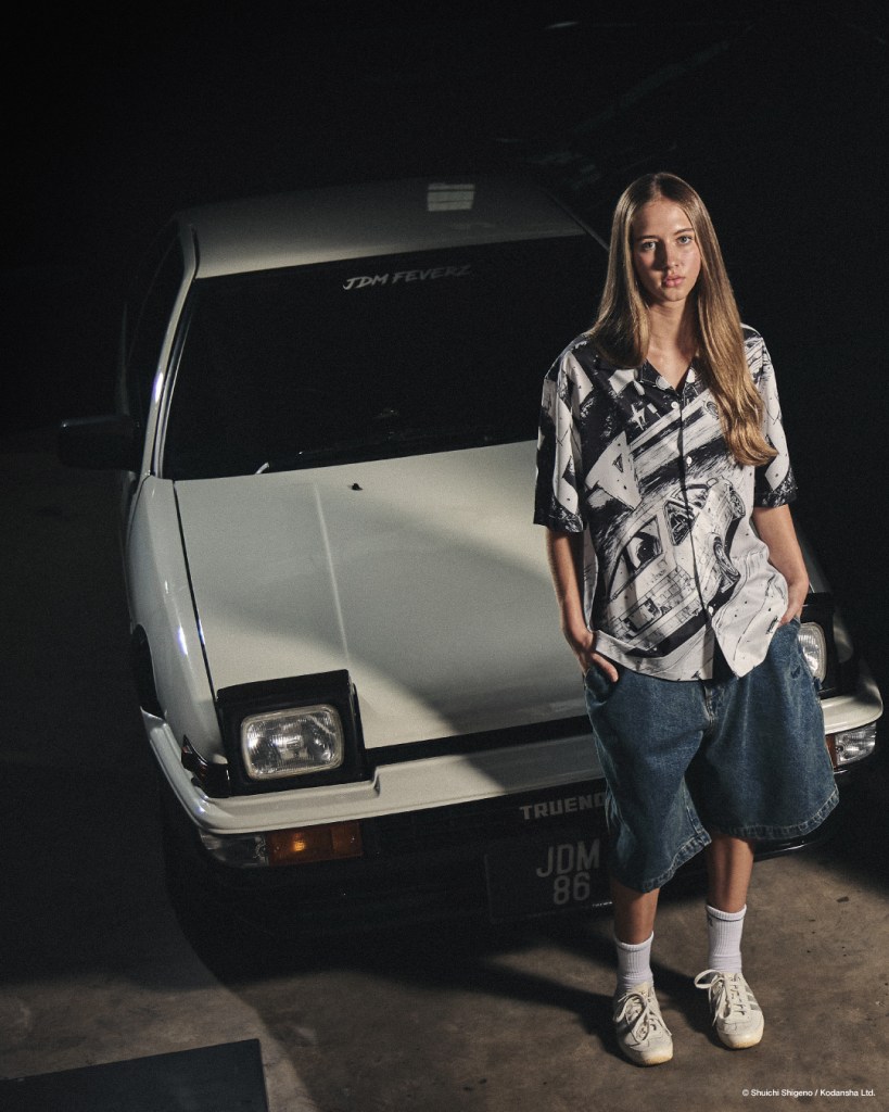 A person wearing a graphic shirt and denim shorts stands next to a classic white car with 'JDM FEVERZ' sticker on the windshield, in a dimly lit setting.