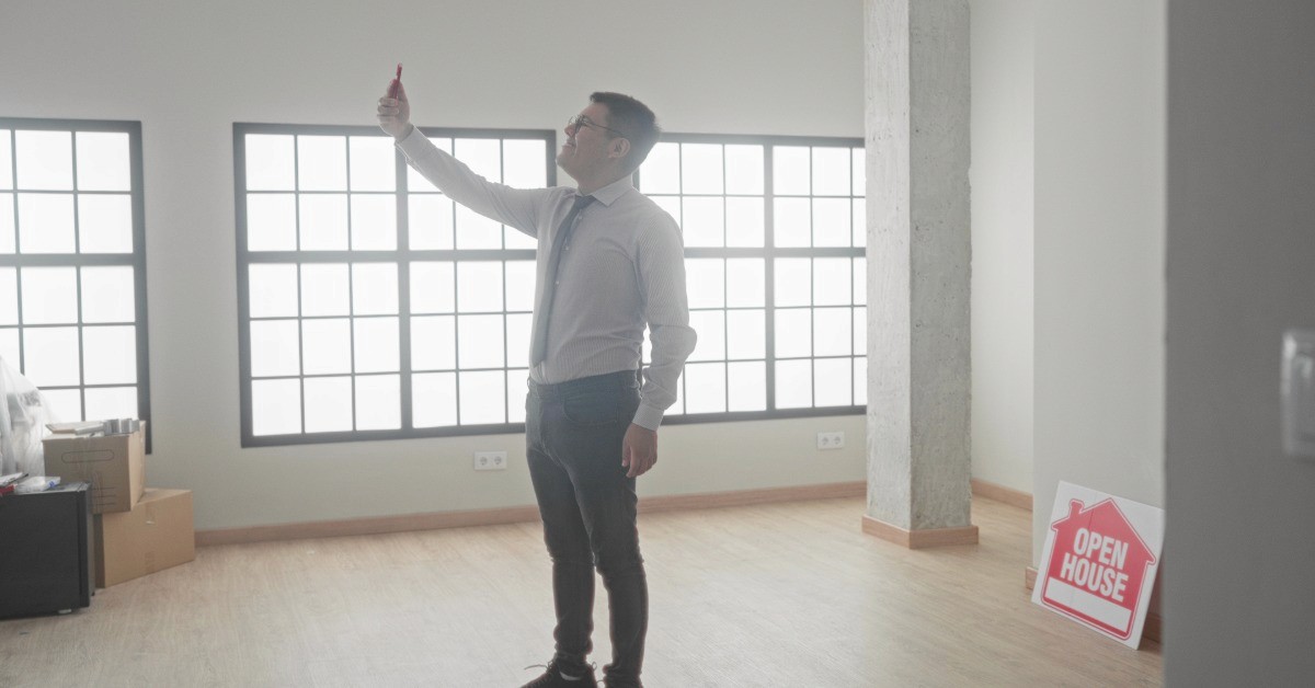 A man wearing a tie stands in an empty home, his phone held above his face. A sign in the corner of the room says "open house."