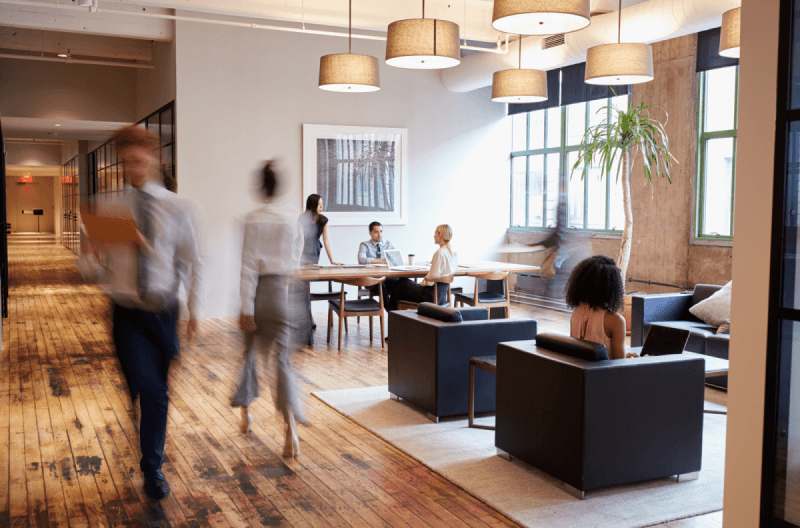 People are working and walking in a modern office space with wooden floors, large windows, and various seating arrangements.