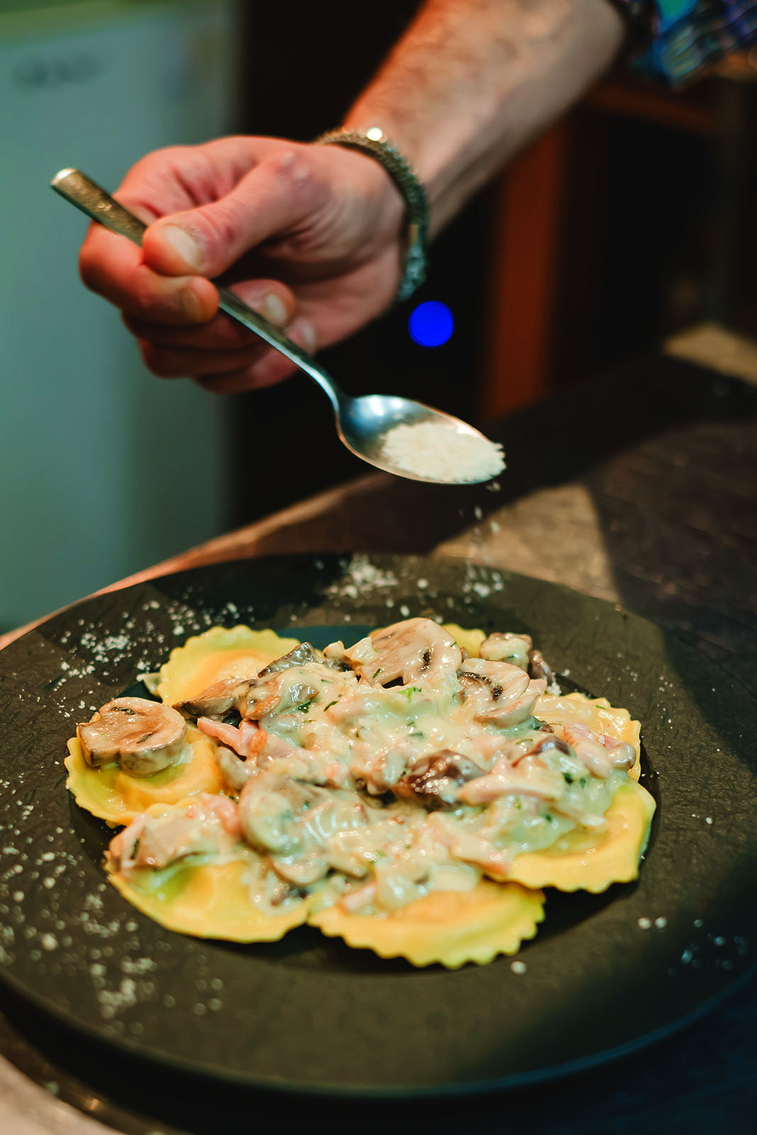 A chef’s hand sprinkles grated cheese from a spoon over a plate of ravioli topped with a creamy mushroom sauce, presented on a dark round plate.