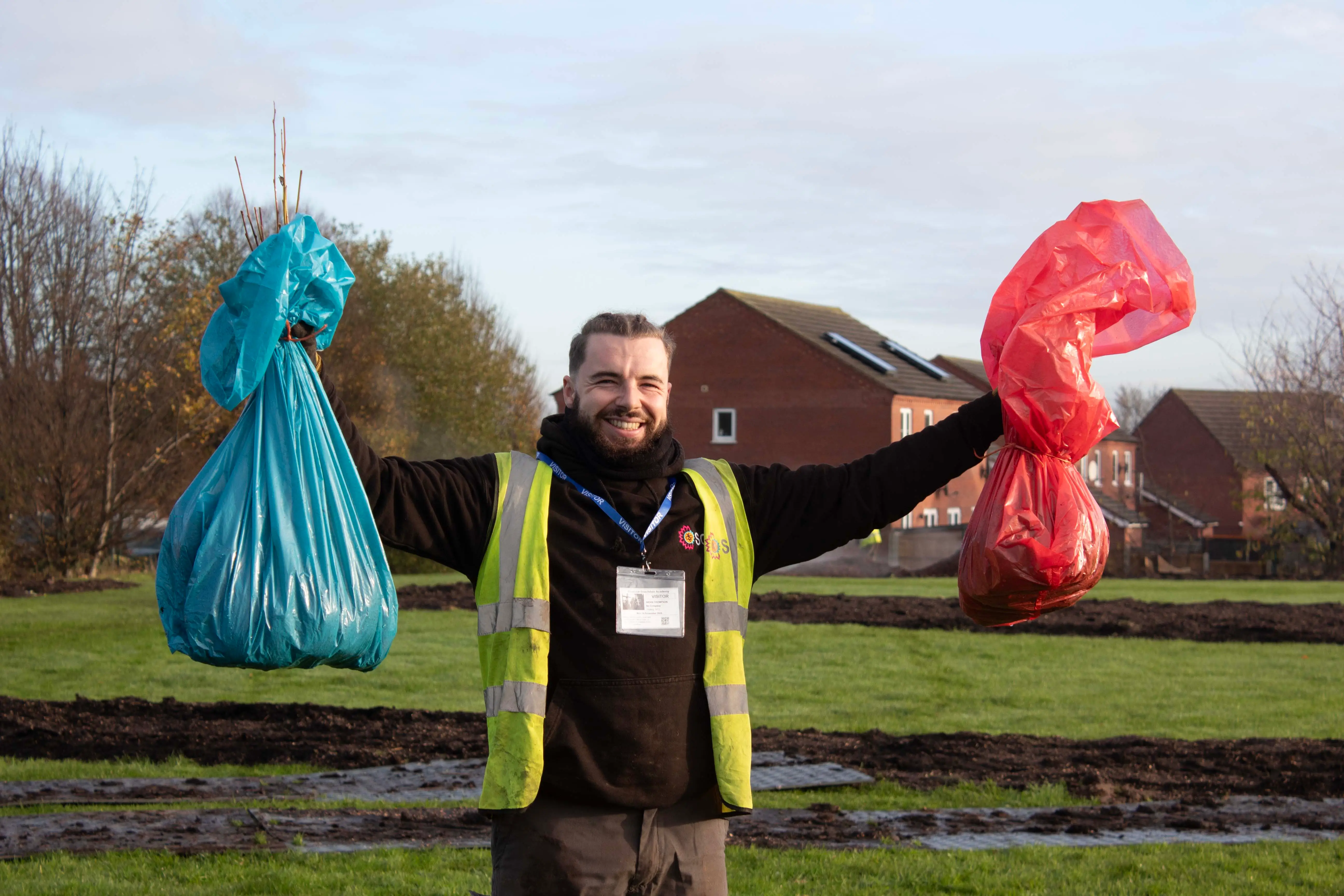 A person in a safety vest holding a blue bag in one hand and a red bag in the other, standing outdoors.