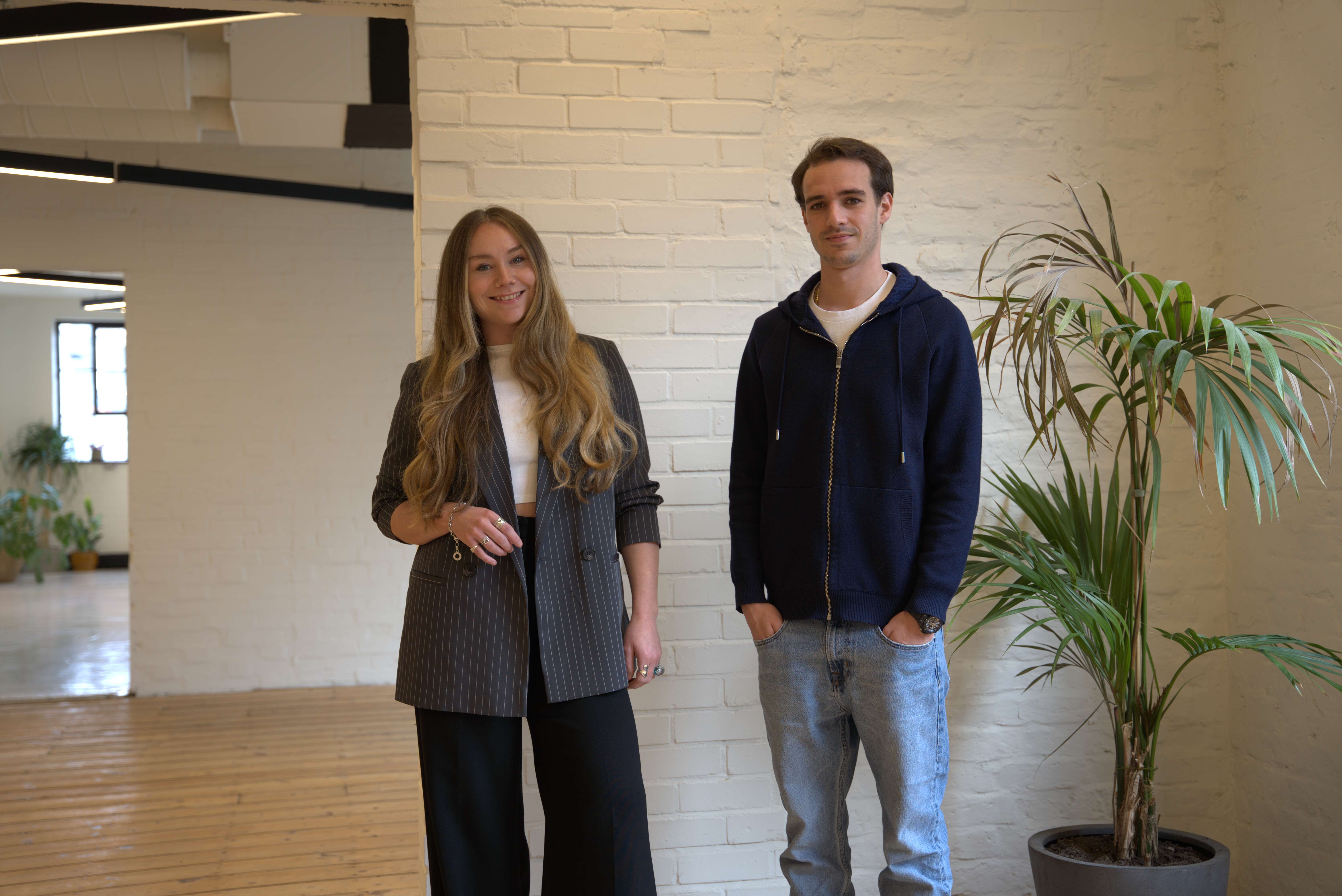 Image of two people - woman and man standing in the centre, looking directly to the camera. Office wall background