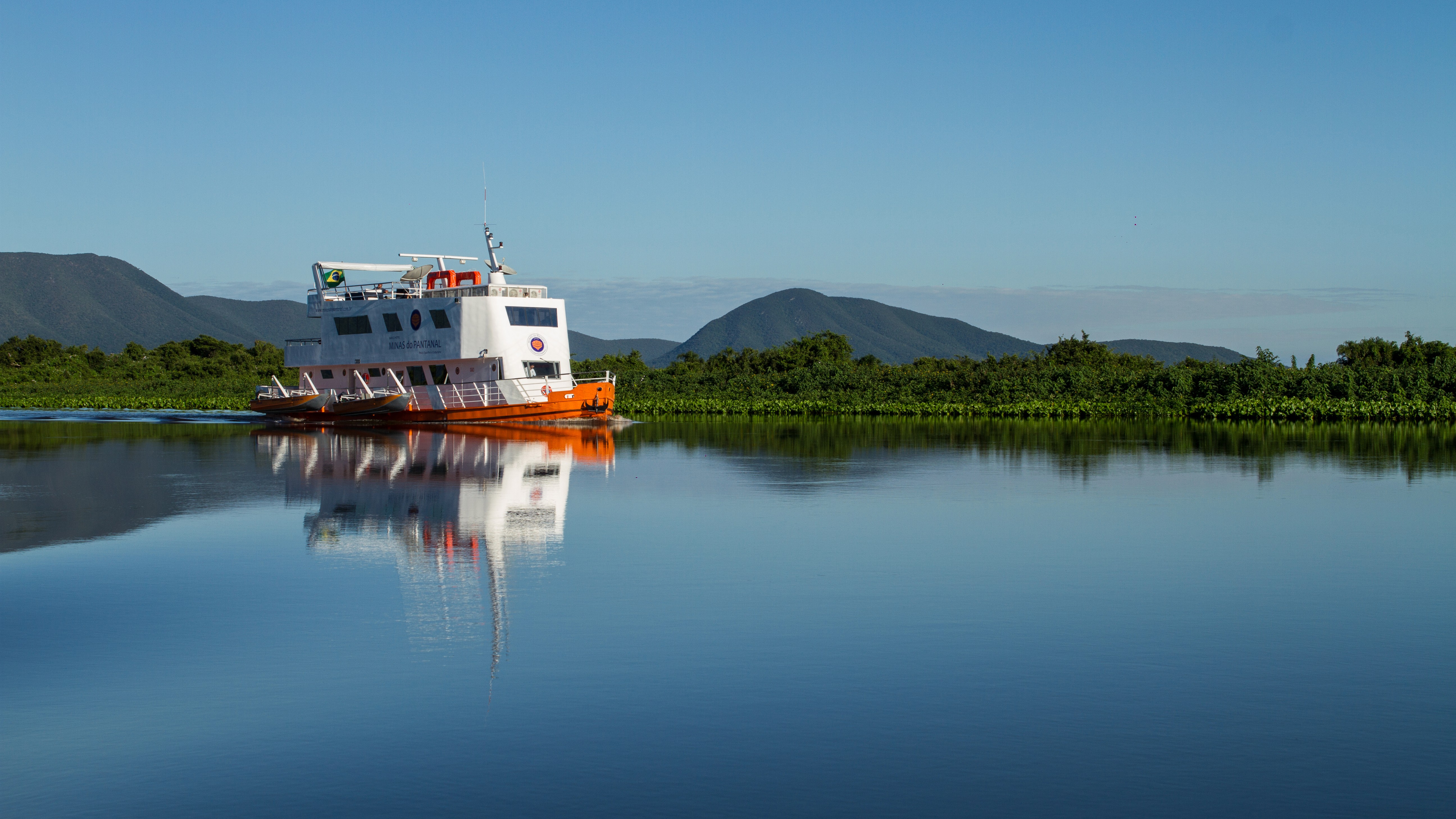 Expedition boat on the Rio Paraguai in the Pantanal, surrounded by lush vegetation and mountains – clear reflection of the vessel on the calm water under a blue sky.