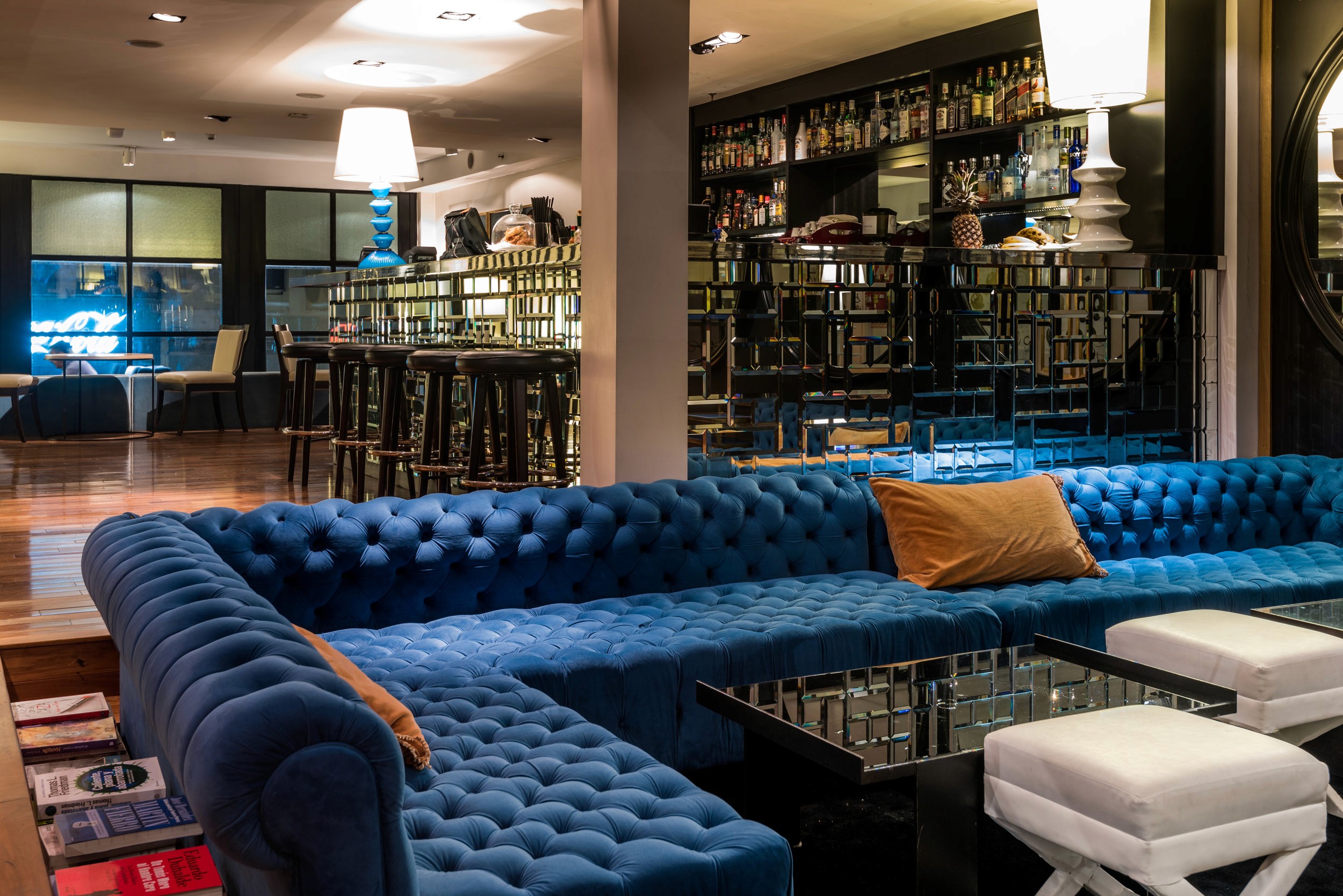 A tufted blue velvet sofa with orange pillows faces a low table in a dimly lit lounge with bookshelves and bar stools.