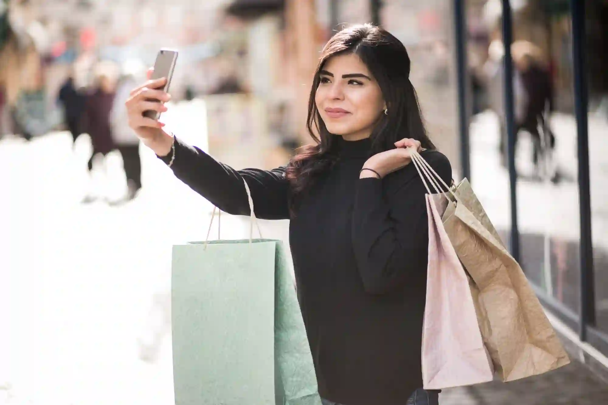 Woman taking a selfie while shopping, posing outside a store window with several brown and green bags.