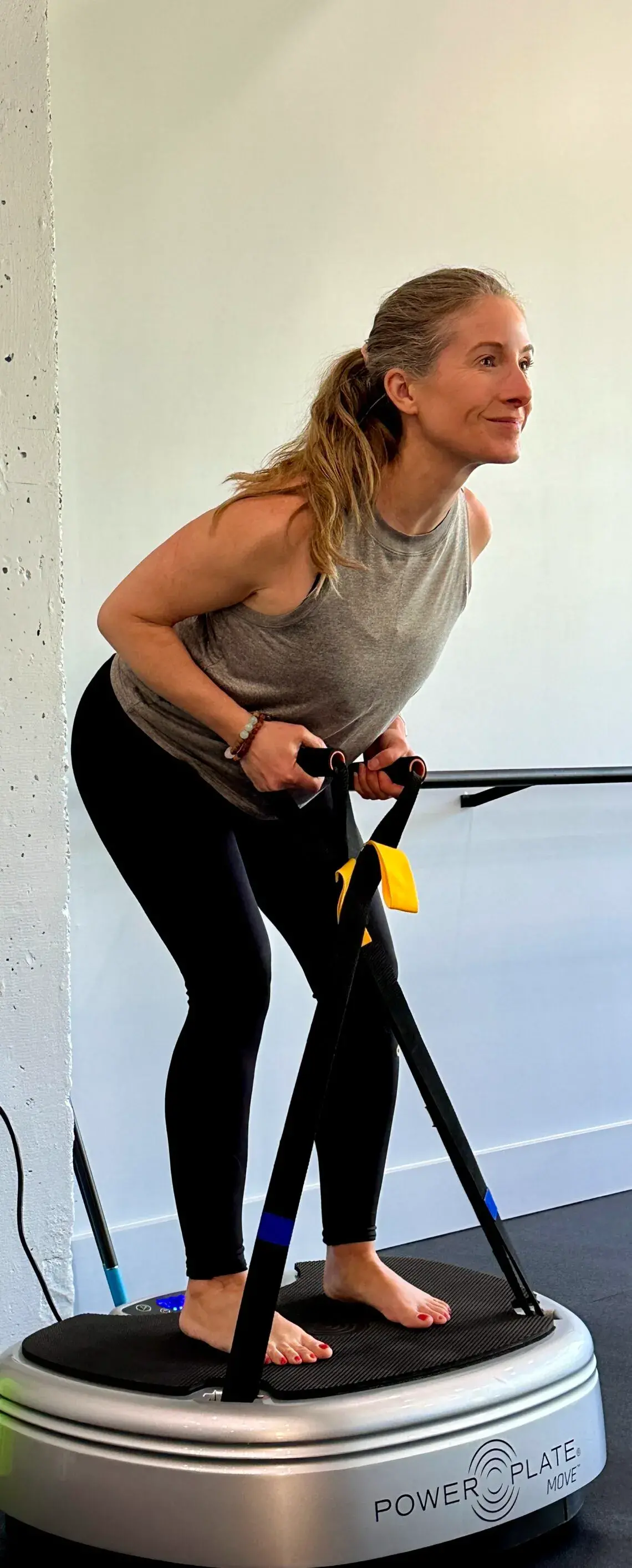 Heather engages in a strength workout on a Power Plate vibration machine, standing barefoot and leaning slightly forward for balance, while holding straps in both hands in a fitness studio setting.