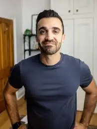 A person with short dark hair and a beard, wearing a fitted gray t-shirt, stands confidently with hands on hips in a room featuring light-colored walls, a wooden door, and a bookshelf.