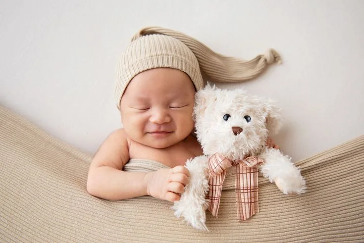 Smiling newborn in a long knit cap beside a white teddy